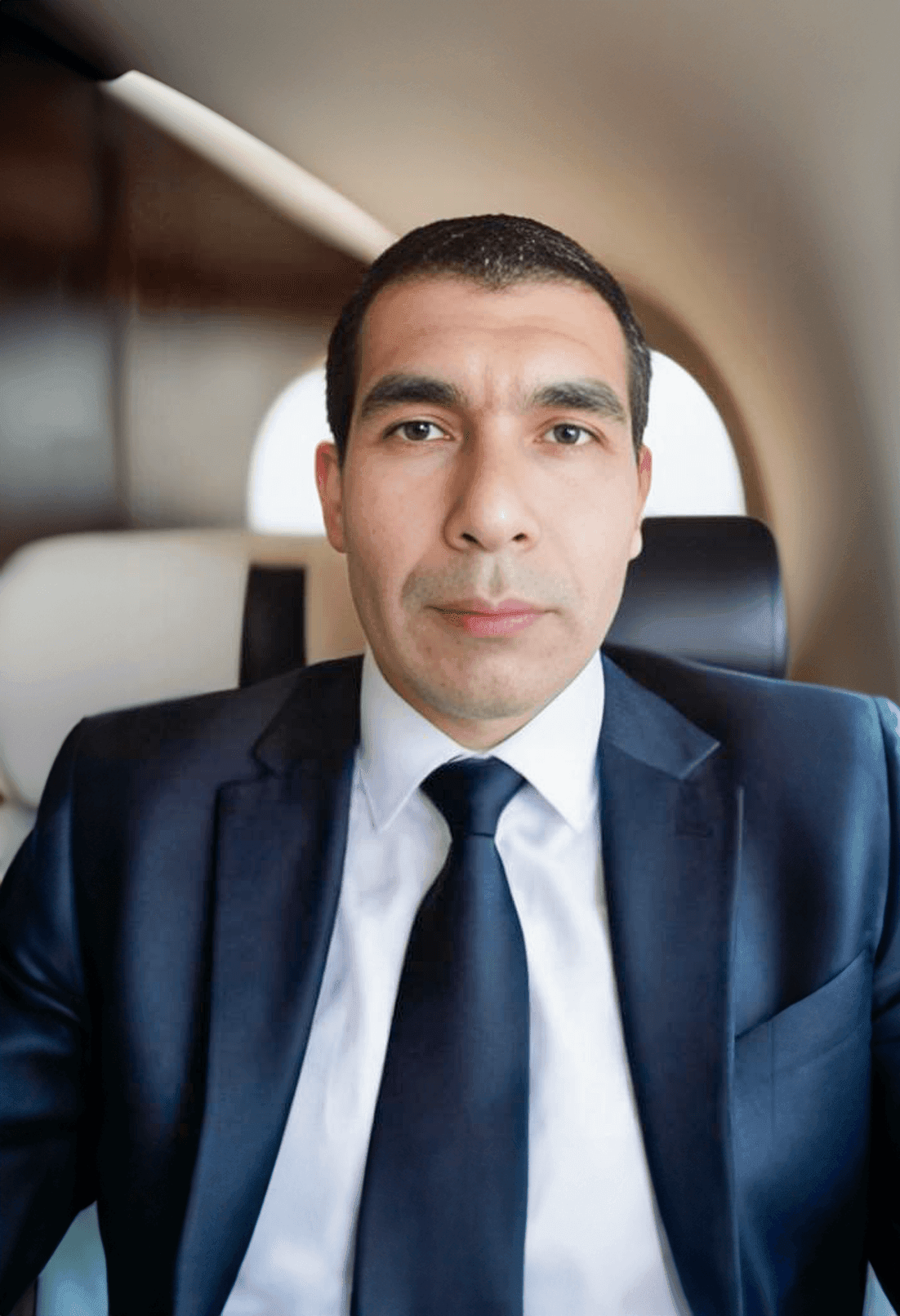 Man in formal suit sitting inside a private jet, looking directly at the camera.